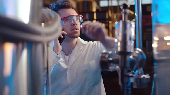Scientist in Lab Coat and Glass Talking on Smartphones Operating Steel Tanks on Panel alt