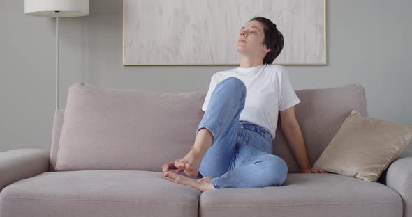 Young Woman in White Tshirt and Jeans Sits on Sofa to Relax Indoors Slow Motion