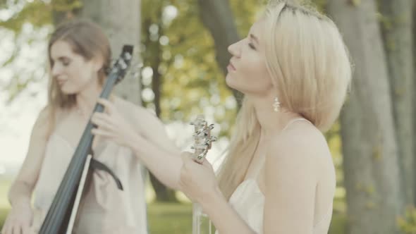 Two Beautiful Girls in Dresses Play the Violin and Cello Standing on the Edge of the Sea Cliff.