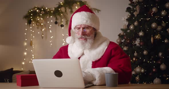 Upset Frustrated Mature Man with Gray Beard in Santa Claus Carnival Costume Looking at Laptop alt