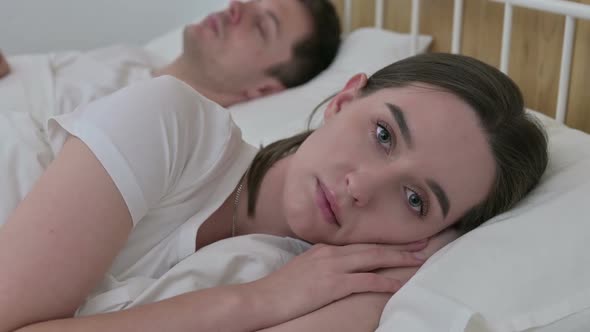 Sad Woman Looking at Camera Next to Sleeping Partner in Bed alt