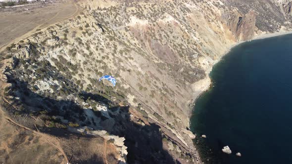 Aerial Drone View of a Man Flying a White and Blue Paraglider Over a Hill and Trees to the Sea Waves alt