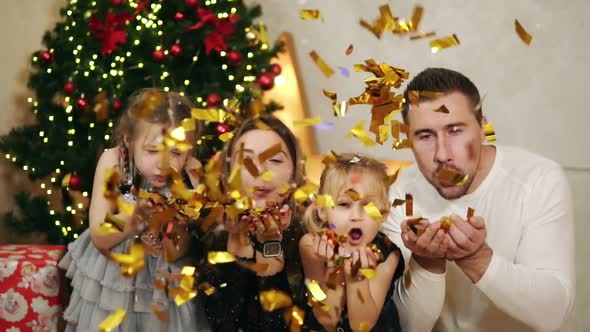 Young Happy Family of Four Sitting By the Christmas Tree and Blowing Golden Confetti alt