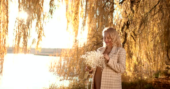 Pregnant Blonde Woman with a Bouquet on the Shore of the Pond alt