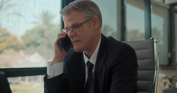 A businessman in formal suit and glasses talking on the phone, smiling, while working at the office. alt