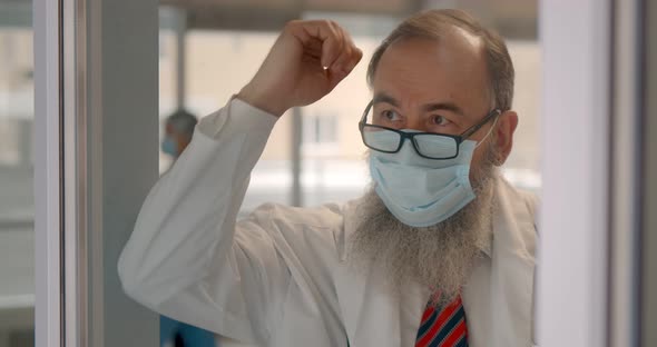 Senior Tired Doctor in Safety Mask Leaning on Glass Door and Relaxing alt