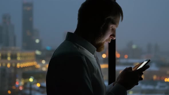 a man uses a smartphone in the dark by the window overlooking the night city