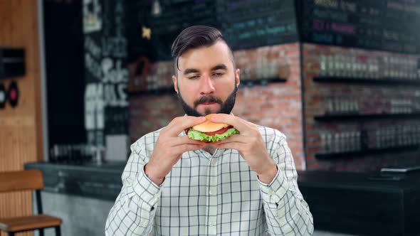 Pleasant Hipster Stylish Young Man Biting Juicy Appetizing Burger Enjoying Taste Looking at Camera alt
