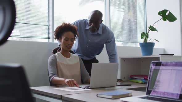 Diverse businessman and businesswoman talking, woman using laptop in office alt