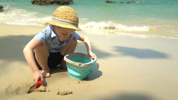 Boy Playing on the Beach By the Sea. He Digs Sand and Throws It in the Bucket alt