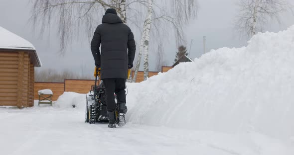 Man Cleans Snow With Snow Plow Background Of Wooden House In Winter alt