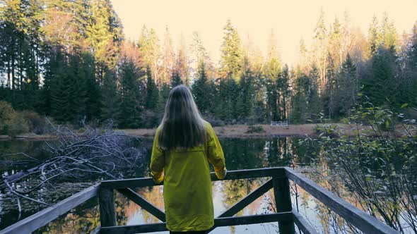 Young Girl in Coat Standing on Terrace Looking at Woodland Landscape in Sunlight alt