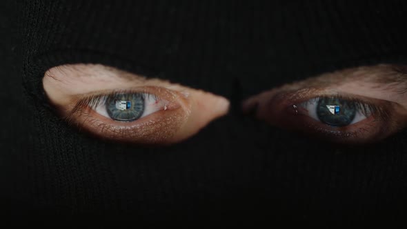Eyes of thief in a black mask (balaclava) before a computer