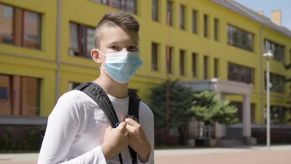 A Caucasian Teenage Boy in a Face Mask Looks at the Camera  a School in the Background