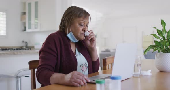 Sick african american senior woman with face mask using laptop at home, with medication on table alt
