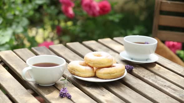 cups of coffee and donuts on a wooden table in a garden with roses on background alt