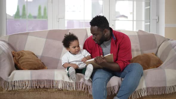 Intelligent Happy Young African American Father Reading Book for Curios Toddler Son Sitting in alt