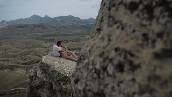A Young Couple Sits on Top of a Cliff Against the Backdrop of Mountains
