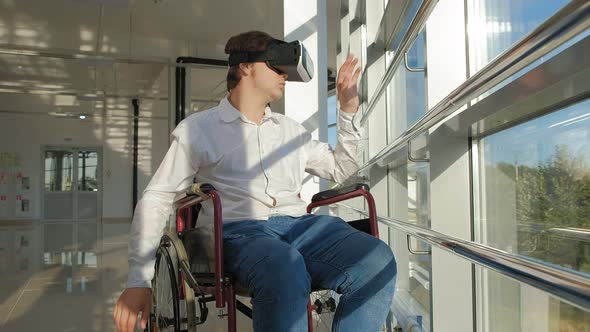 Disabled Man on a Wheelchair at a Window Uses a Helmet of Virtual Reality