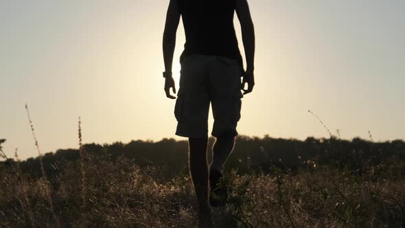 Silhouette of Young Man Walking in Field To Setting Sun and Raising Hands Up alt