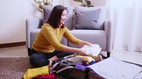Asian woman arranging clothes in takeout suitcase in her living room. alt