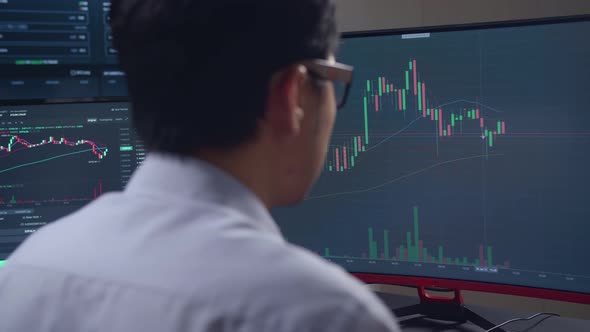 Asian Male Stock Market Broker Looking Analyzing Graphs On Multiple Computer Screens alt