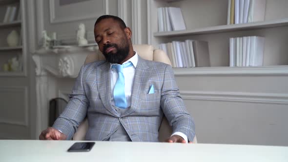 a Bearded AfricanAmerican Man in a Plaid Suit with a Blue Tie is Sitting at a Table on a Light Chair alt