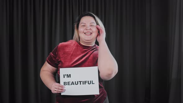 A Confident Flirty Fat Woman Holding a Nameplate with a Sign I'M BEAUTIFUL alt