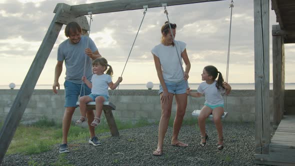 Happy Family Rolls Young Children on a Swing Outdoors at Sunset alt