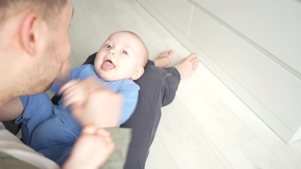 Father Plays with a Smiling Baby Son Lying on Knees at Home Together