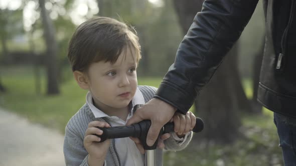 Joyful Caucasian Little Boy Riding Scooter in Sunlight with Young Male Hand Pulling Steering Wheel alt