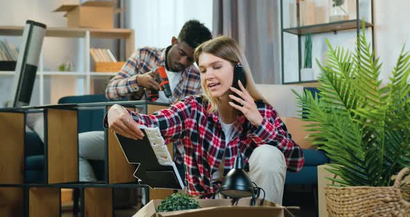 Girl Unpacking Cartons During Phone Conversation while Her Positive Skilful Black-Skinned Boyfriend alt