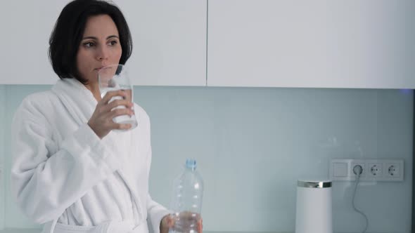 Thirsty smiling 35s woman in white bathrobe drinking water