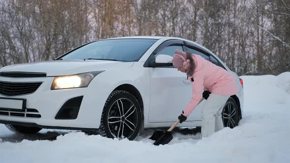 Female Digging Snow with Shovel From Wheel of Car alt