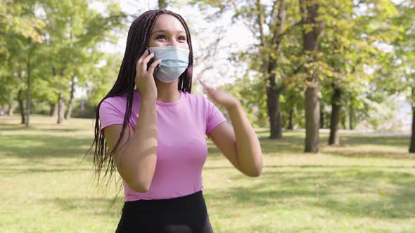 A Young Black Woman in a Face Mask Talks on a Smartphone, Excited, in a Park on a Sunny Day alt