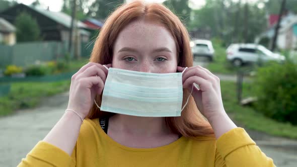 Redhead Girl with Freckles Puts on a Medical Mask While Standing on the Street alt