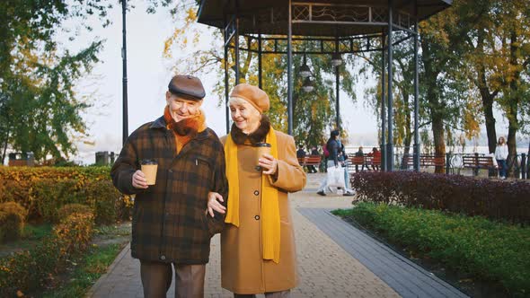 Happy Couple Grandparents Holding Paper Cups of Coffee Talking and Smiling During a Walk in Autumn alt