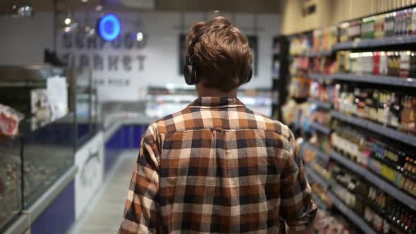 Stylish Caucasian Guy with Headphones Walks Through Goods Section of the Store Wearing Plaid Shirt alt