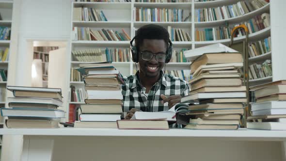 Happy African Male Student Listening Music while Sitting at the Table in the Library alt