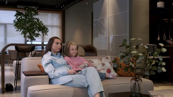 Happy young woman is sitting on cozy sofa with adorable little cute daughter. Watching TV alt