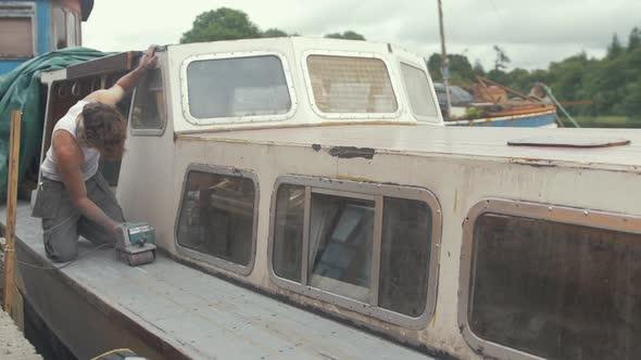 Young working topless in Summer sealing wooden boat cabin roof to prevent leaks. alt