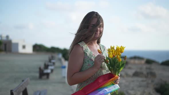 Portrait of Romantic Young Caucasian LGBT Woman with Bouquet of Flowers and Rainbow Flag Standing on alt