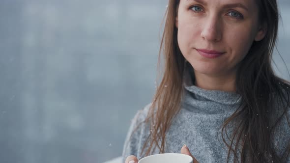 Caucasian Woman Stays on Balcony During Snowfall with Cup of Hot Coffee or Tea alt