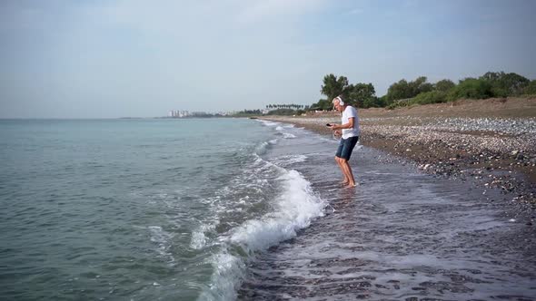 A Slender Whitehaired Elderly Man in White Headphones is Dancing Energetically Barefoot on the Beach