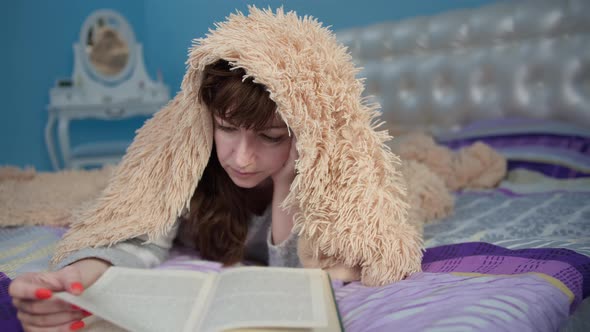 Brunette Woman Covered with a Blanket Lies on the Bed and Reads a Book