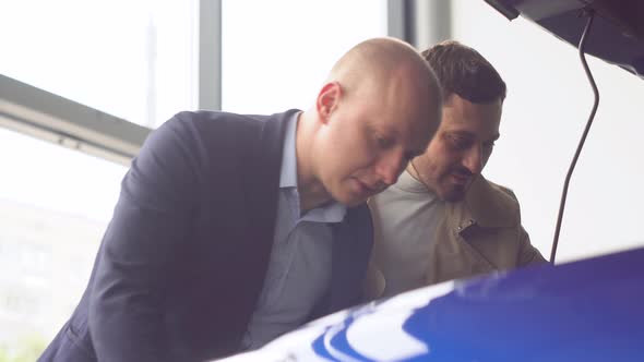 Professional Salesman Smiling Showing a New Car at the Dealership to His Male Customer alt