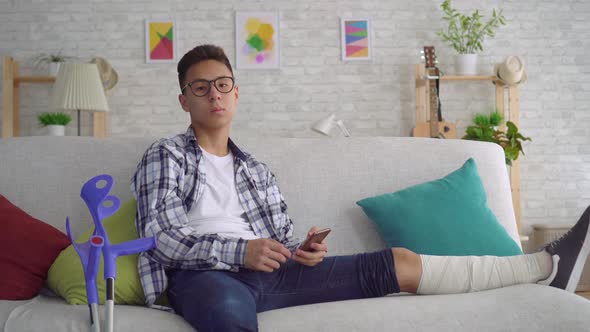 Sad Asian Young Man with a Broken Leg in Bandages Lying on the Couch Uses Smartphone and Looking at alt