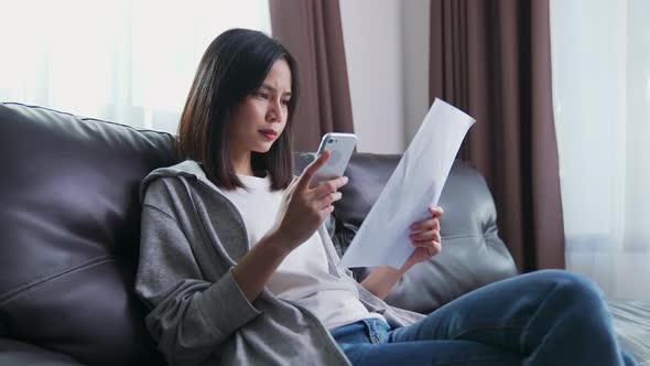 Asian woman holding the bill and smartphone in the living room and stressed by excessive expenses. alt