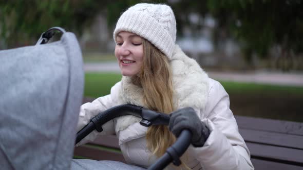 Young Mother with Newborn Child Outdoor. She Sitting on the Bench with Baby Sleeping in Pram alt