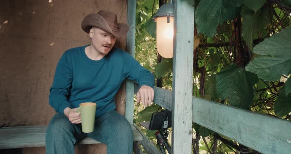 Cowboy Man on Porch Drinks Coffee From a Mug and Communicates Online By Phone